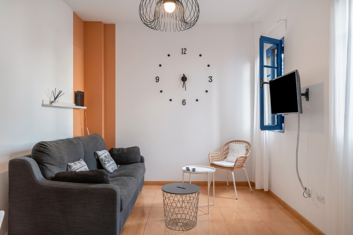 A comfortable living area is presented with a dark grey sofa adorned with decorative cushions. A wall clock with a minimalist design is featured, and a round white coffee table is positioned in front of the sofa. A rattan chair is included, with a television mounted on the wall nearby.