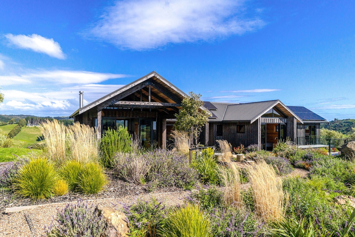 A modern lodge is set amidst carefully landscaped gardens with lush greenery and native plants. The building features a combination of wooden and metal elements, with expansive windows that invite natural light. The blue sky and distant hills form a scenic backdrop.
