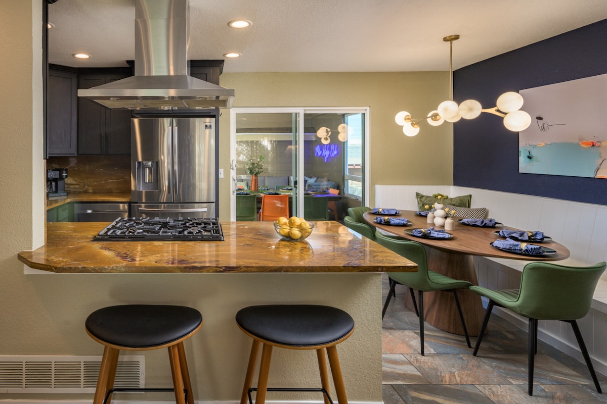 The open kitchen features modern appliances and a smooth countertop, with bar seating for two. A spacious dining area is visible, showcasing a wooden table set for eight, illuminated by stylish pendant lighting, plus vibrant blue decor and green chairs.