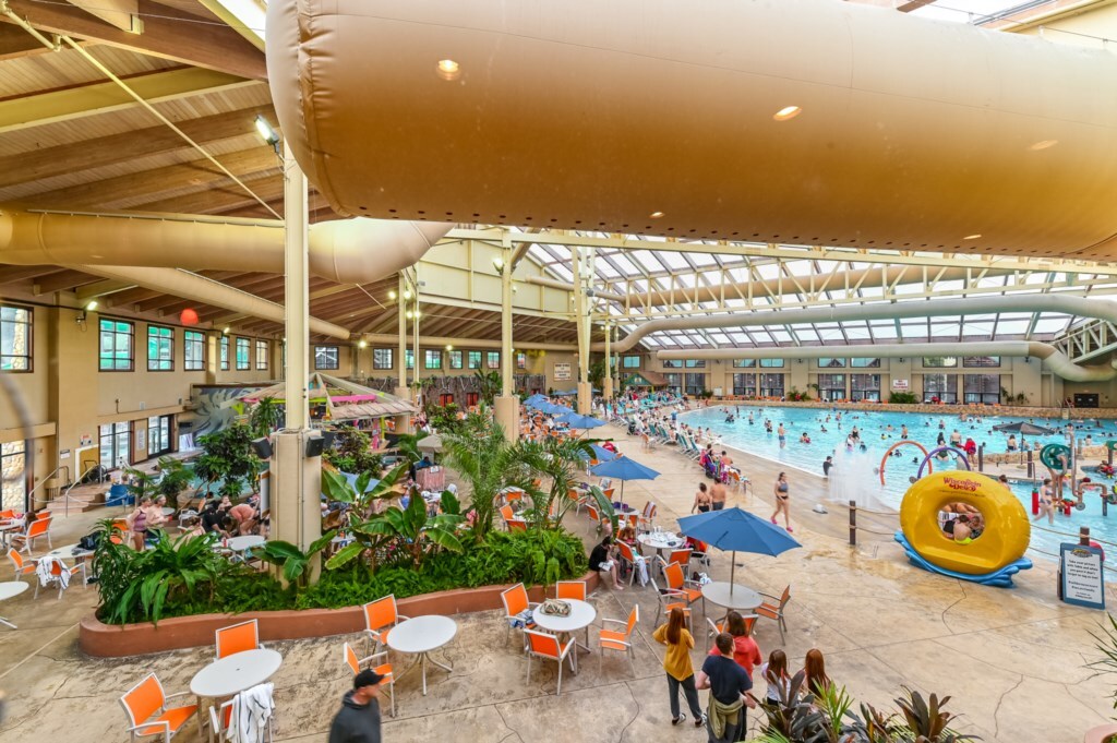 An indoor water park is vibrant with families enjoying various water activities. A large pool is surrounded by colorful lounge chairs and umbrellas, while lush greenery adds natural accents. Ample seating areas are available for relaxation, providing a lively atmosphere for recreation.