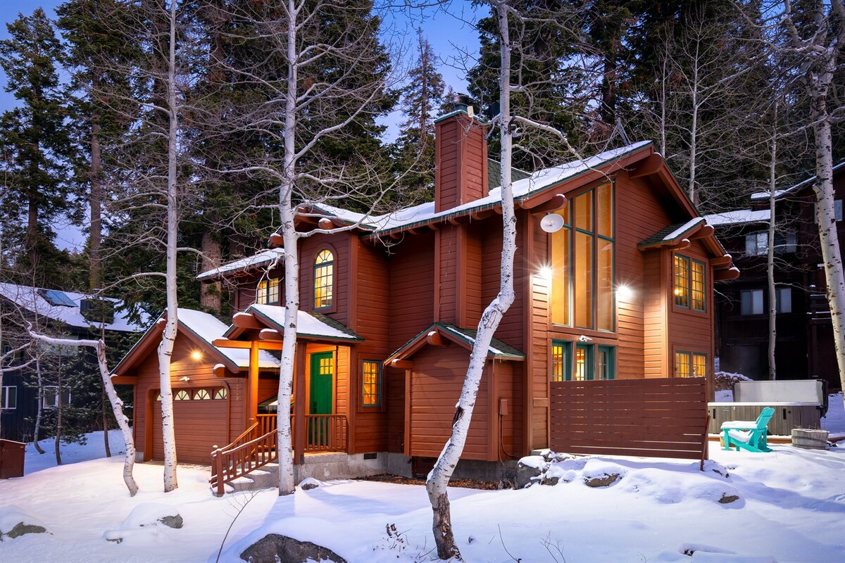 The cabin's exterior combines wood and stone materials, surrounded by snow-covered ground and trees. Large windows and a green door enhance the inviting character, and the structure is complemented by a garage and wooden stairs leading to the entrance.