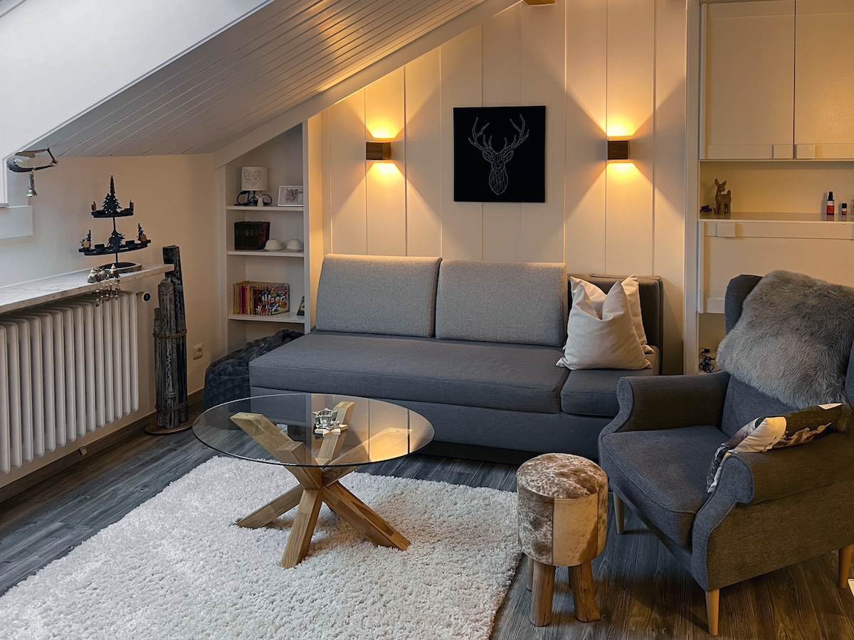 A stylish living area is presented, featuring a gray sectional sofa and a cozy armchair. A glass coffee table sits atop a plush white rug. Soft lighting from the wall sconces illuminates a mounted deer head art piece above the couch, enhancing the room's inviting feel.