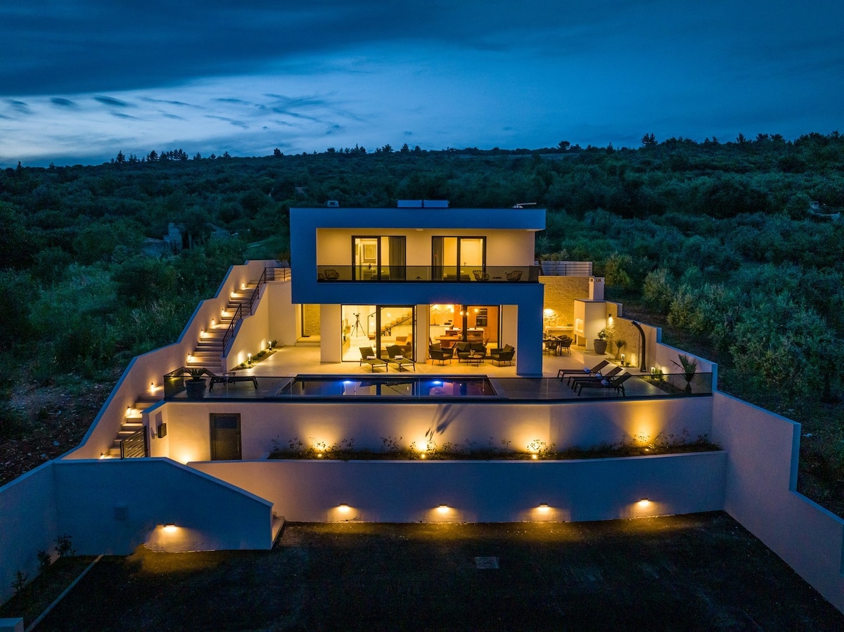 The villa is illuminated at night, showcasing a modern design and multiple levels. Soft lighting highlights the exterior and terrace areas, while expansive glass doors reveal a cozy indoor space, inviting relaxation in the evening ambiance.