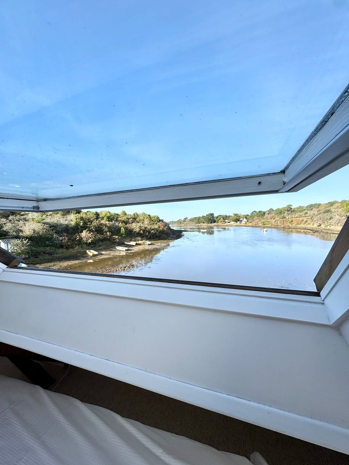 A window is seen open, revealing a scenic view of a tranquil river framed by lush greenery. Bright blue skies reflect on the water's surface, enhancing the serene atmosphere of the outdoor landscape.