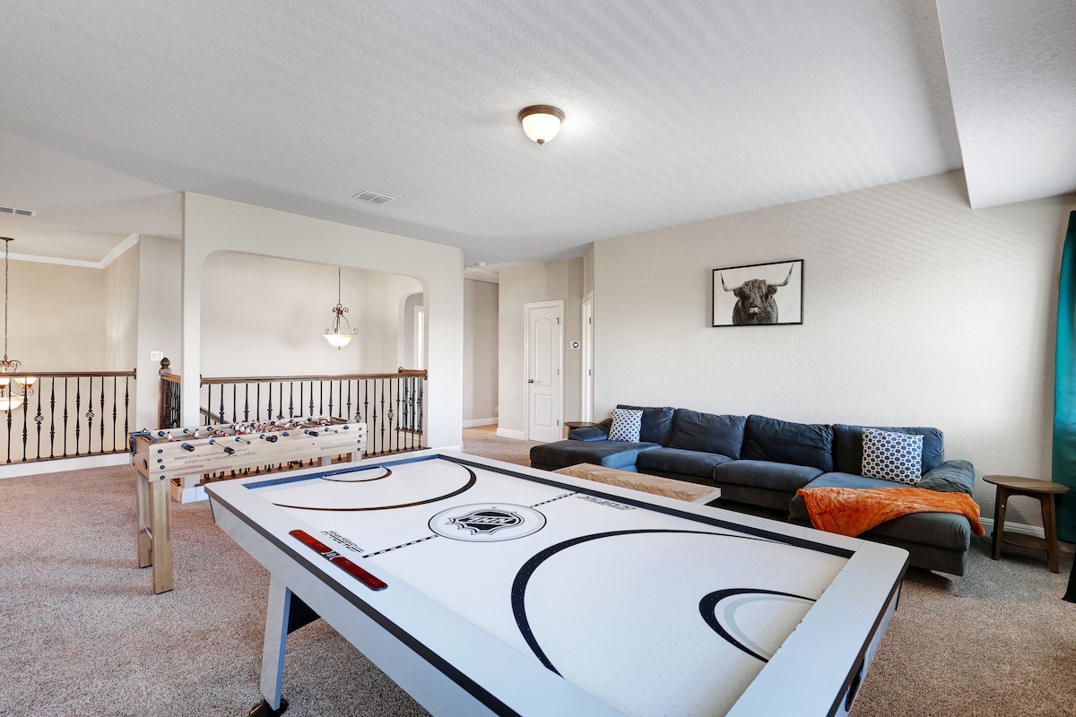 A spacious game room features a large air hockey table and a comfortable sectional sofa. Soft lighting enhances the inviting atmosphere, while a vibrant orange throw blanket adds a pop of color to the seating area.
