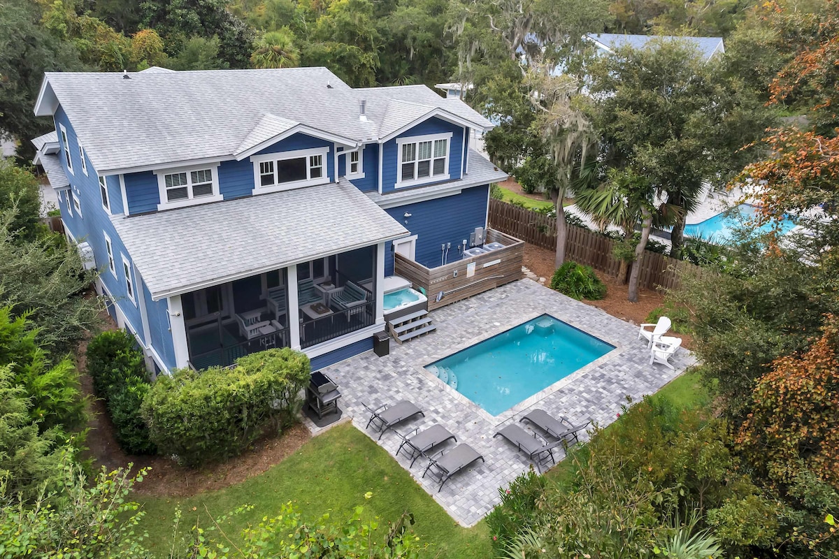An aerial view showcases a spacious blue house with a light grey roof, surrounded by lush greenery. A private pool is set in a paved patio area, accompanied by several lounge chairs. The outdoor deck adjacent to the house offers additional seating.