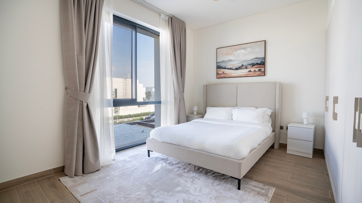 A serene bedroom is furnished with a spacious bed, complemented by two nightstands with lamps. Natural light filters through a large window, offering views of the outdoor area. Soft, neutral tones and light textures create a calming ambiance.