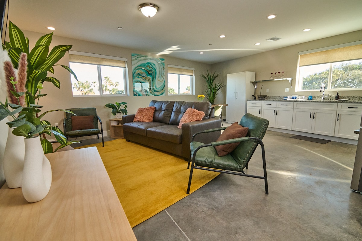 An open-concept living area features a comfortable dark sofa and two green chairs. A bright yellow area rug contrasts with light grey floors. Natural light floods in through large windows, highlighting a modern kitchen against a vibrant wall art piece, complemented by indoor plants.