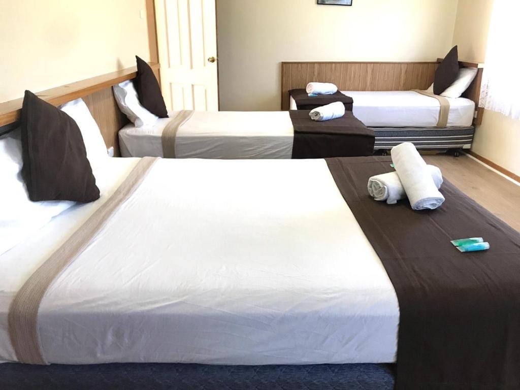 The sleeping area features two spacious beds, each made with neutral-toned linens and dark throw pillows. A third bed, positioned to the side, complements the arrangement. Natural light filters through a window, illuminating the simple décor and organized space.