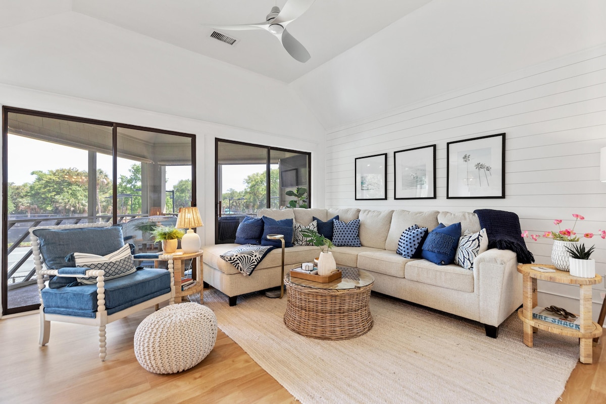 A comfortable living room features a spacious sectional sofa adorned with various cushions, a soft pouf, and a round coffee table made of woven material. Natural light floods in through the large windows, providing views of the outdoor space. Framed artwork is displayed on the wall.