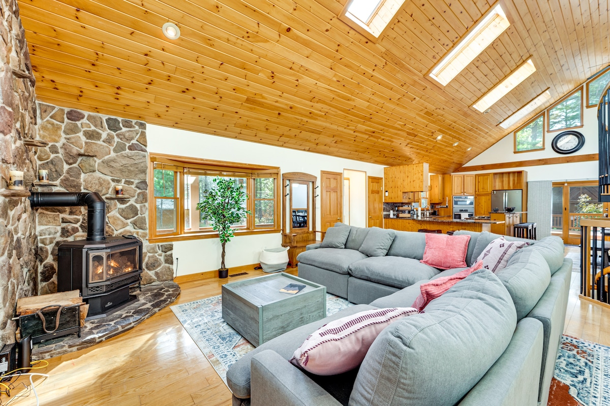 The open living area features a stone fireplace, surrounded by soft seating and a large coffee table. Skylights allow natural light to brighten the wood-paneled ceiling, complementing the spacious layout. Natural greenery is present, enhancing the inviting atmosphere.