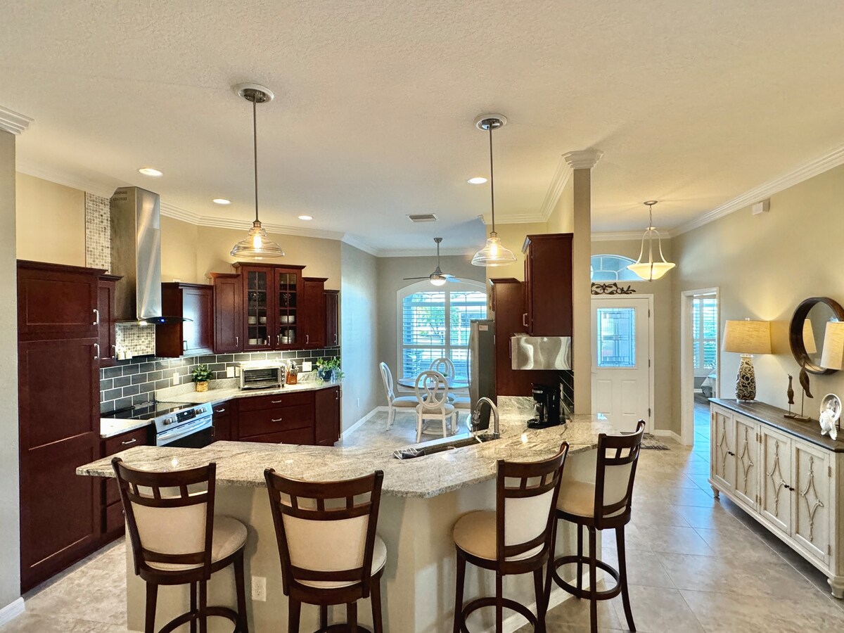 An open-concept kitchen features dark wood cabinetry and a spacious countertop with four bar stools. Modern appliances and a tile backsplash are visible. Natural light fills the area, highlighting the adjacent dining space with a round table and additional seating. Stylish fixtures add to the ambiance.