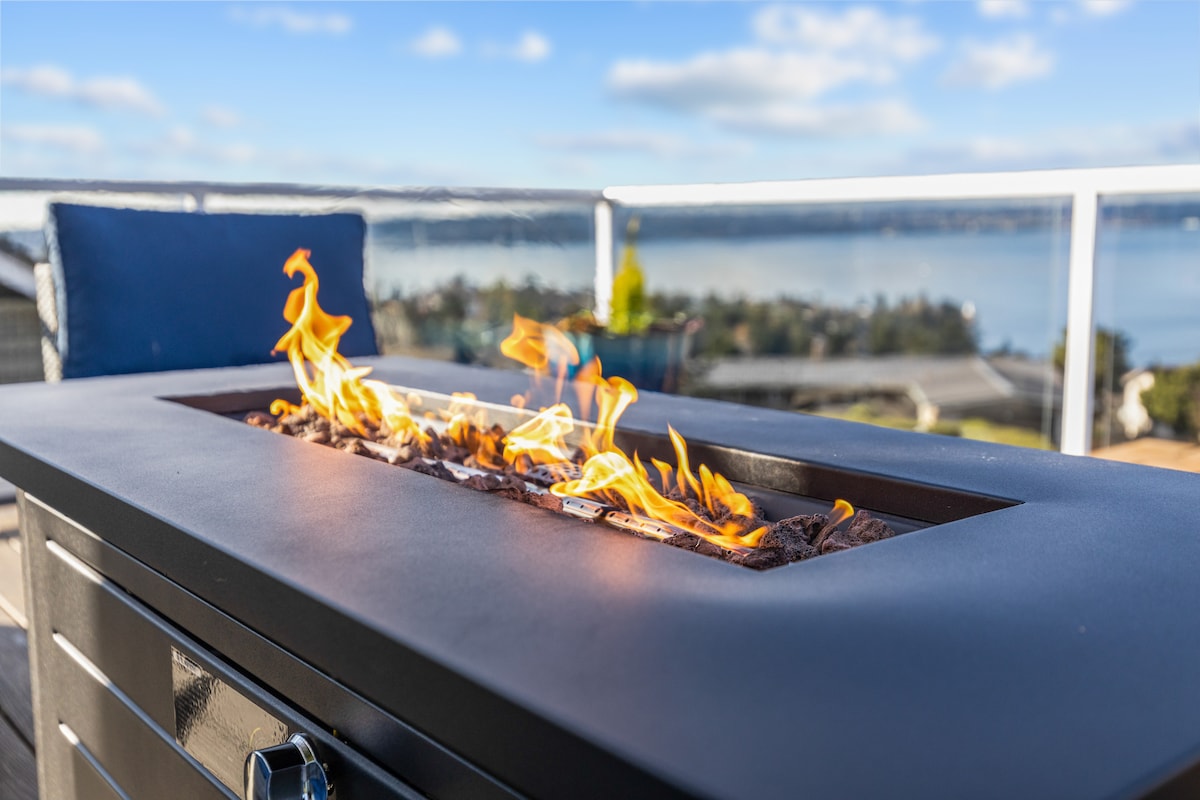 An outdoor gas fire pit is centered on a deck, featuring flickering flames over decorative stones. The backdrop showcases a scenic view of a tranquil waterfront and distant hills under a clear sky, creating a serene atmosphere for relaxation.