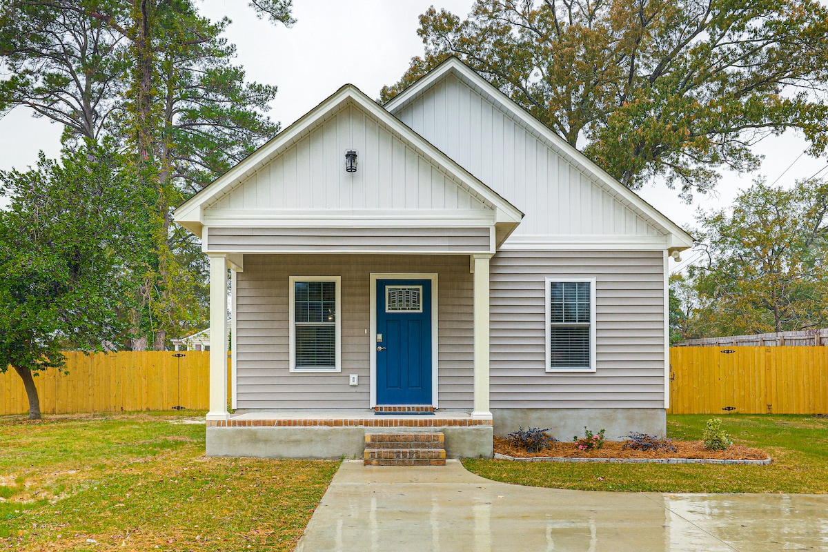 The exterior of a single-story home is presented, featuring a light gray facade and a bold blue front door. The porch is supported by two square columns, and the driveway is visible, leading to a smooth, concrete surface. A fenced yard is seen in the background, complemented by greenery.