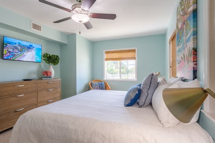 Prostrana spavaća soba s velikim bračnim krevetom, komodom, televizorom i udobnom stolicom od ratana. Uređenje uključuje tropsku umjetnost, zidove u svijetloplavoj boji i tkanine na prozorima, što stvara svijetlu i opuštenu obalnu atmosferu.