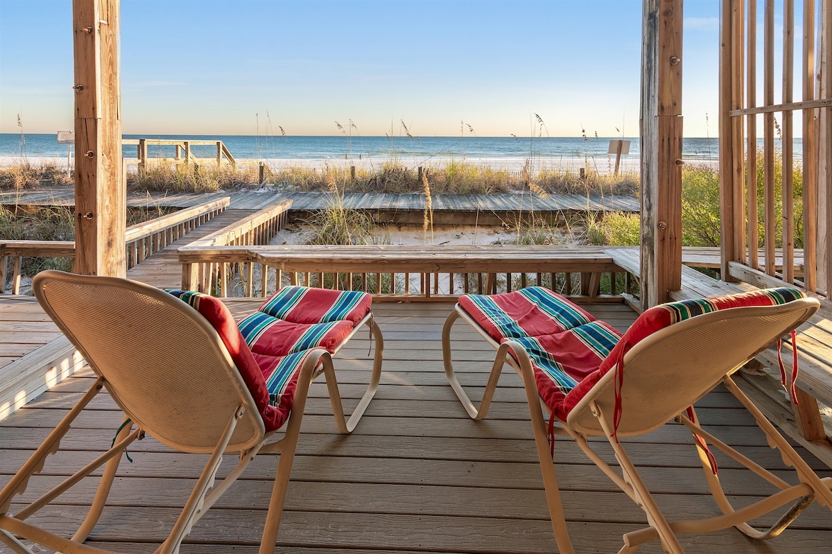 Two lounge chairs with colorful cushions are positioned on a wooden deck, overlooking a sandy beach and the Gulf of Mexico. Soft grasses sway in the gentle breeze, while a tranquil blue sky merges with the horizon, enhancing the serene coastal atmosphere.