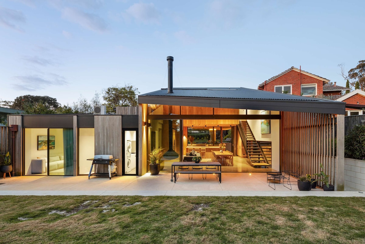 The exterior of a modern architectural home is showcased, featuring large glass windows that open to a spacious outdoor area. A wooden deck with seating is visible, alongside a landscaped lawn. The home's design incorporates natural materials and a sleek black roof.