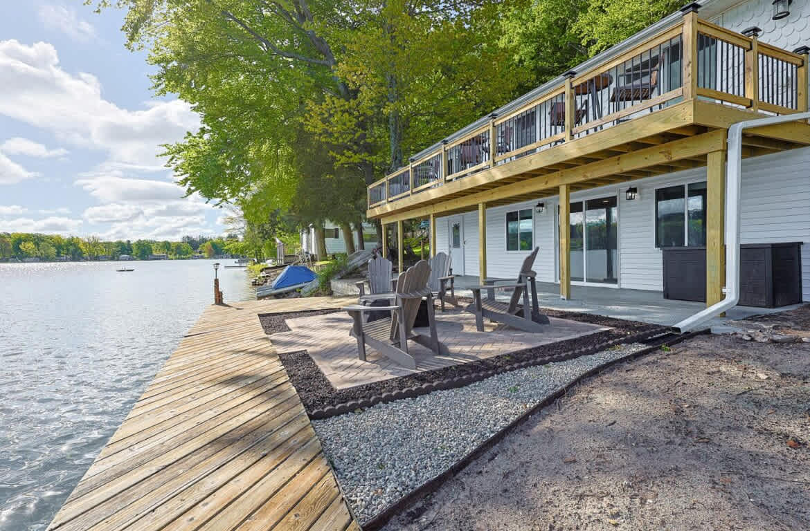 A wooden dock extends alongside Crystal Lake, with Adirondack chairs positioned on a natural surface. The home's expansive deck above features railings, allowing for views of the water. Lush green trees offer shade, and sunlight reflects off the serene lake surface.