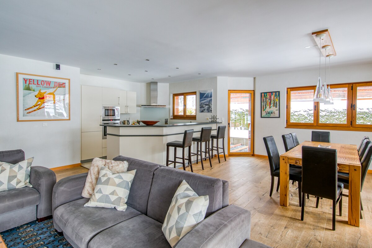 An inviting living area features a spacious gray sofa arranged with patterned cushions. A dining table for eight is positioned near large windows, providing natural light. The kitchen bar is equipped with high chairs, while artistic prints adorn the walls, enhancing the modern decor.
