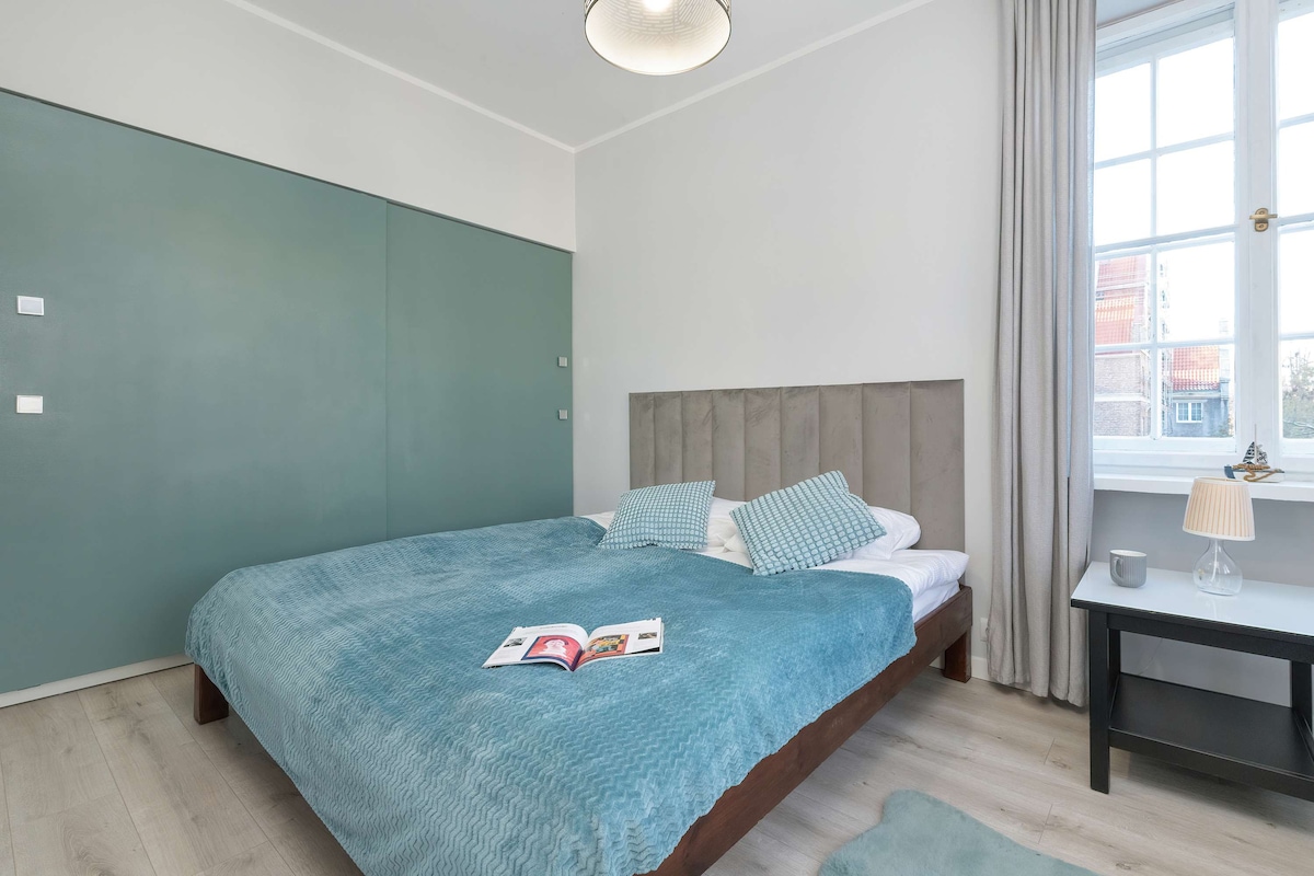 Chumba cha kulala kilicho na ukuta wa kijani kibichi, kitanda chenye matandiko mawili na madirisha makubwa yanayoruhusu mwanga wa asili.