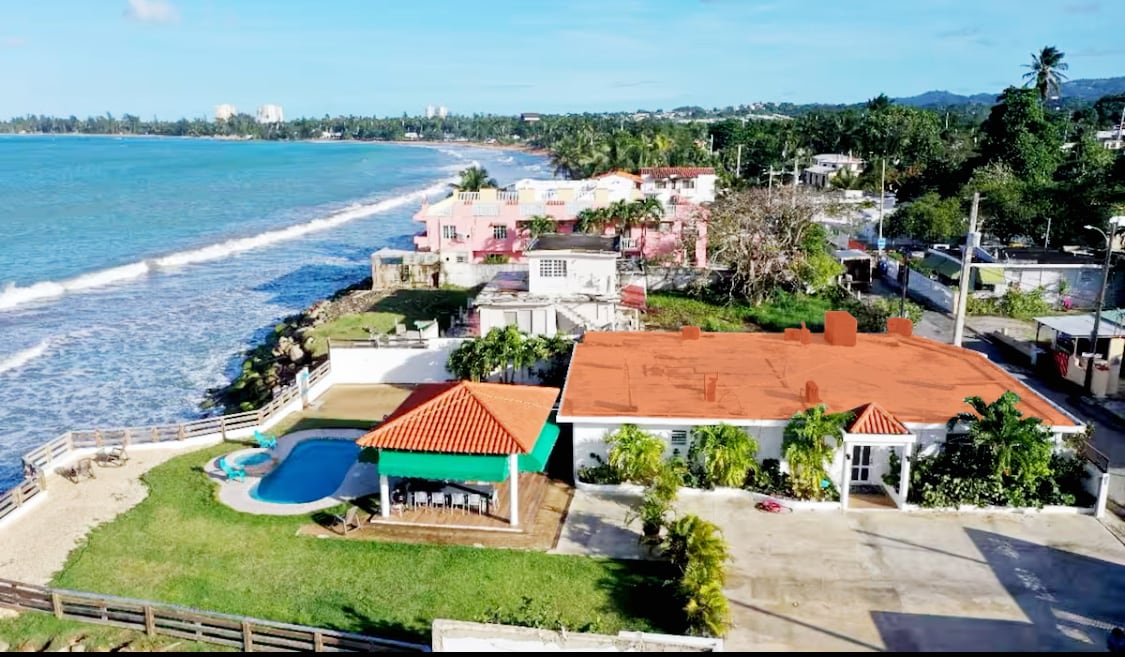 An aerial view captures the spacious beachfront home with a swimming pool and a vibrant orange-roofed structure. Adjacent properties with varied colors are visible, and the ocean waves gently lap at the shoreline. Lush greenery frames the outdoor living space.