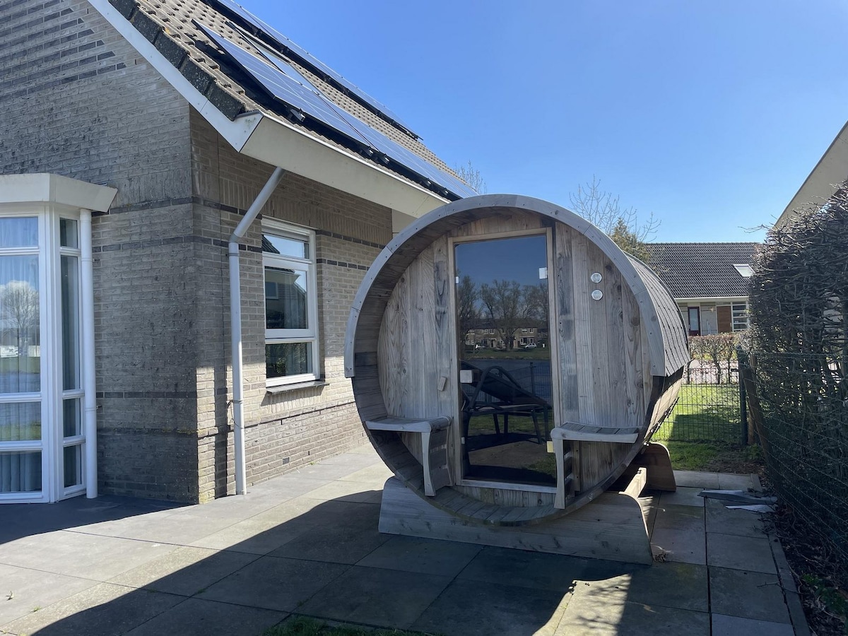 A wooden infrared sauna with a circular design is placed beside the bungalow on a paved area. Two lounge chairs are positioned in front of the sauna, surrounded by greenery and a clear sky. The exterior of the bungalow is visible, showcasing a blend of stone and wood.