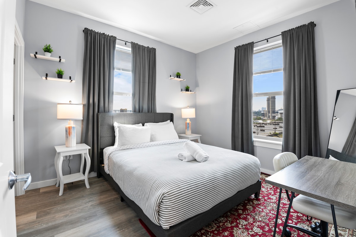 A well-appointed bedroom features a spacious bed with crisp bedding and neatly folded towels. Two bedside tables with lamps flank the bed, while shelves with small plants are mounted on the wall. A desk is placed beside a window dressed with light-filtering curtains.