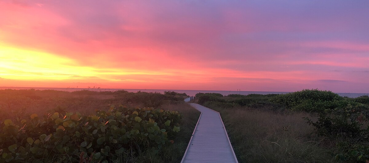 A wooden boardwalk leads through lush greenery towards the beach, with soft grasses on either side. The sky is filled with vibrant hues of pink and orange from a setting sun, creating a serene backdrop over the tranquil sea.