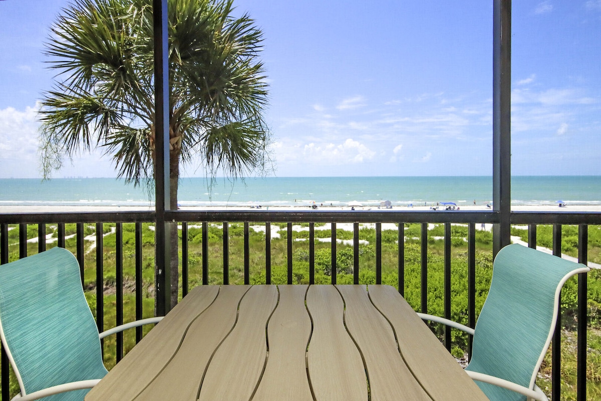 A private balcony features a light, wooden table surrounded by two turquoise chairs. The vibrant green palm tree is visible, framing a panoramic view of the ocean and beach, under a bright blue sky with scattered clouds.