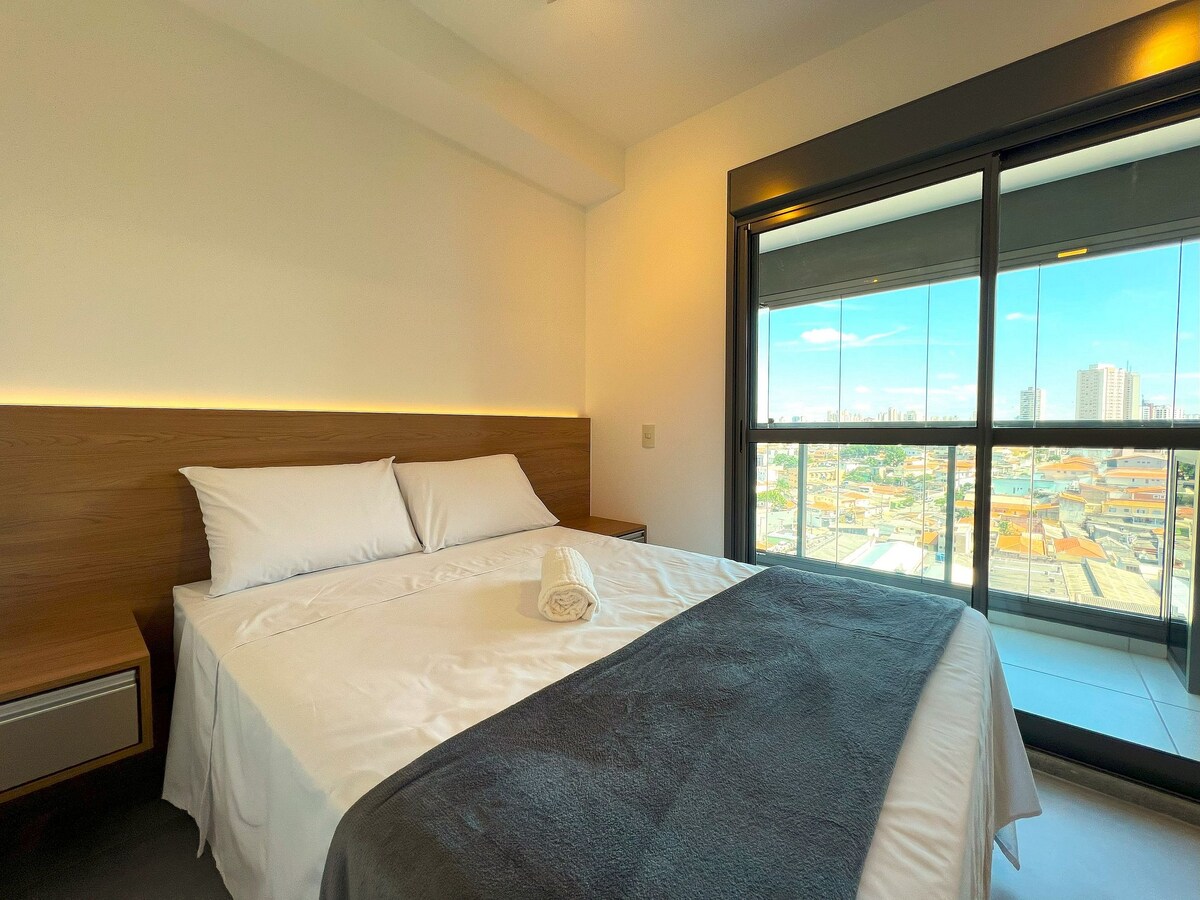 A modern bedroom features a spacious bed with a light-colored duvet and a dark throw. Large glass doors provide a view of the cityscape, allowing natural light to fill the room. Soft lighting is accentuated along the headboard.