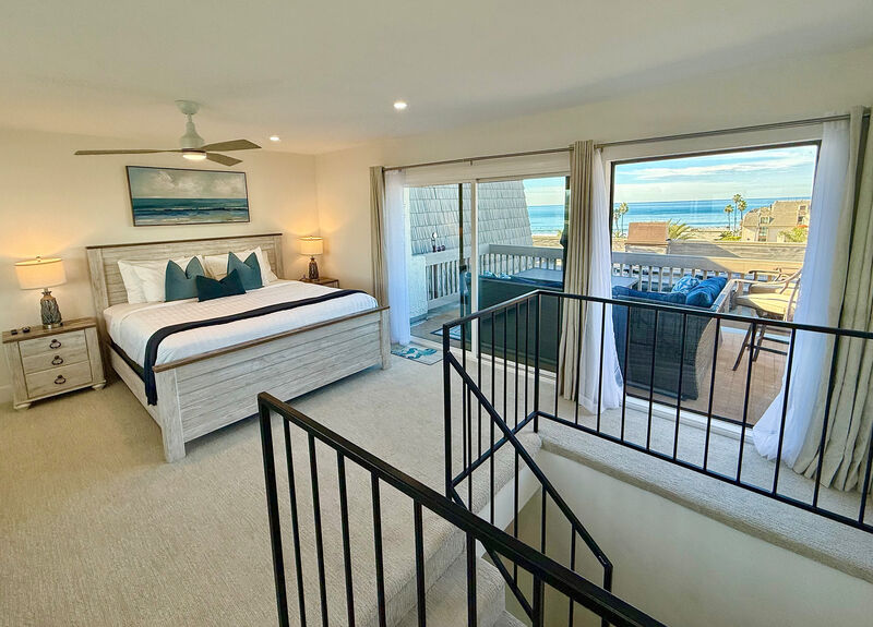 A spacious bedroom features a king bed with layered bedding and decorative pillows. Two large windows frame ocean views, while a ceiling fan provides airflow. A bedside table and modern lamp complement the decor, with a staircase leading to the sunlit upper deck.