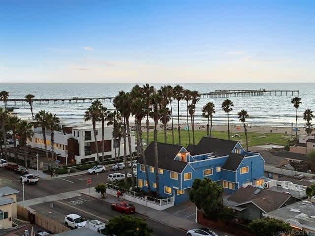 An elevated view captures a vibrant blue building framed by palm trees, positioned near the beach. The ocean stretches into the distance, with gentle waves lapping at the shore. A pier extends into the water, completing the coastal scene.