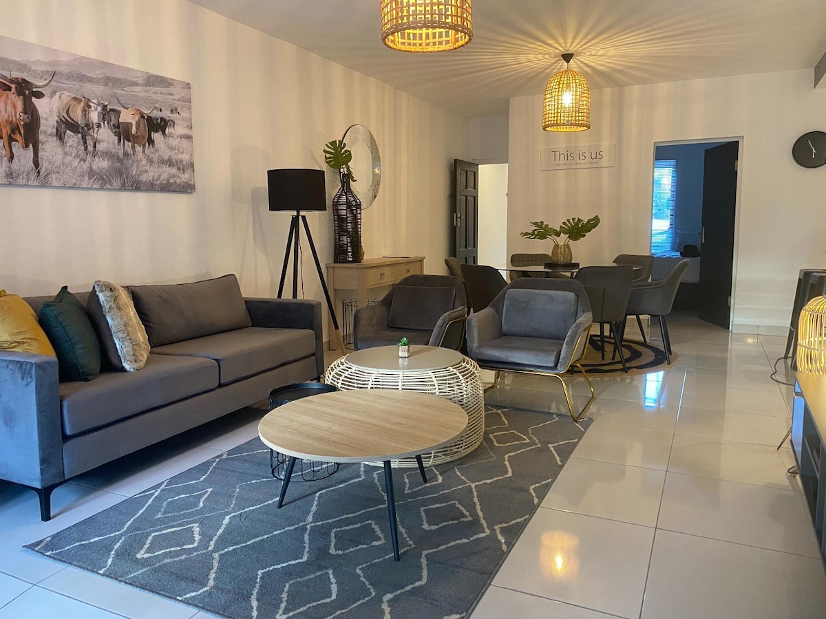 A modern living area features a mix of comfortable seating, including a plush sofa and stylish chairs, arranged around a wooden coffee table. Light fixtures hang from the ceiling, casting a warm glow. A decorative rug lies on the glossy tiled floor.