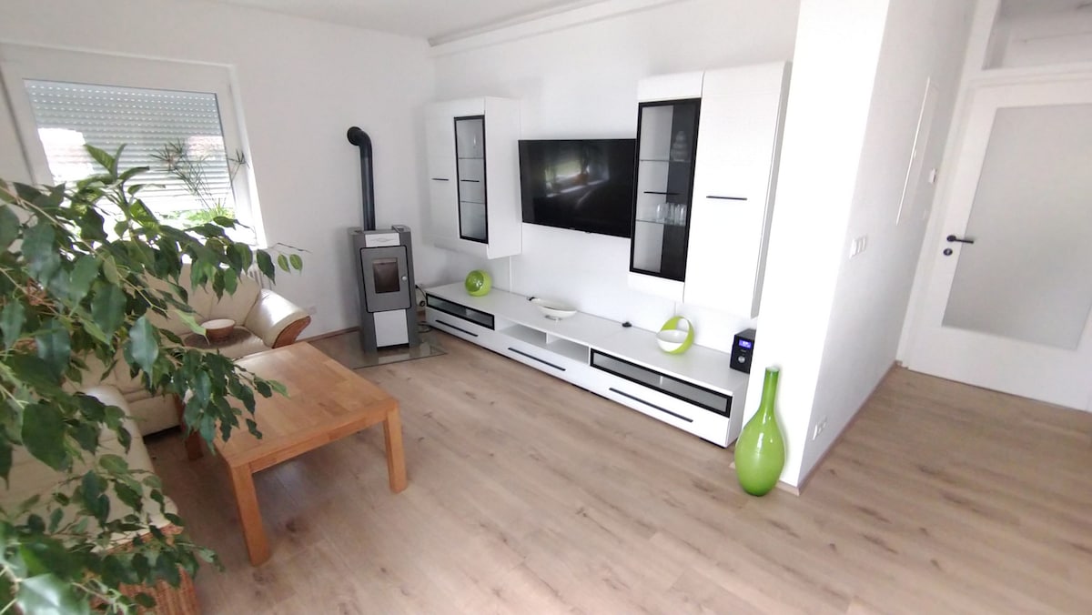 A modern living room features a light wood floor and sleek white furniture. A flat-screen TV is mounted on a wall unit, accented by decorative vases in green. A comfortable sofa is positioned near a large window, allowing natural light to fill the space.
