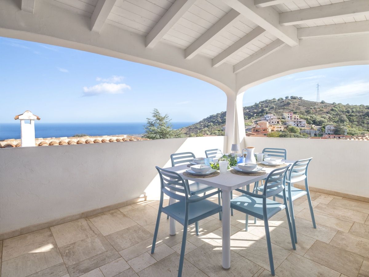 A dining area features a white table surrounded by six light blue chairs, positioned under a covered terrace. A panoramic view of the sea and hills is visible in the background, enhancing the outdoor ambiance.