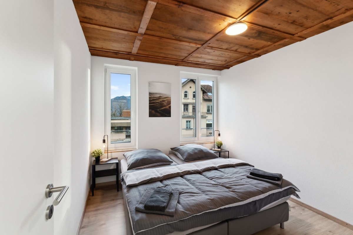 A cozy bedroom is presented with two comfortable single beds positioned together, dressed in fresh linens. Natural light enters through two large windows, highlighting the wooden ceiling. Nightstands with reading lamps are placed on either side of the beds, adding functionality to the space.