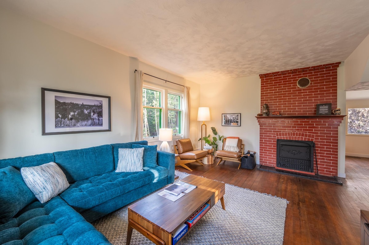 A cozy living room features a large, plush teal sectional sofa paired with two warm wood armchairs. A rustic coffee table sits on a textured area rug, while a brick fireplace adds a charming focal point. Natural light streams through the windows, enhancing the inviting atmosphere.