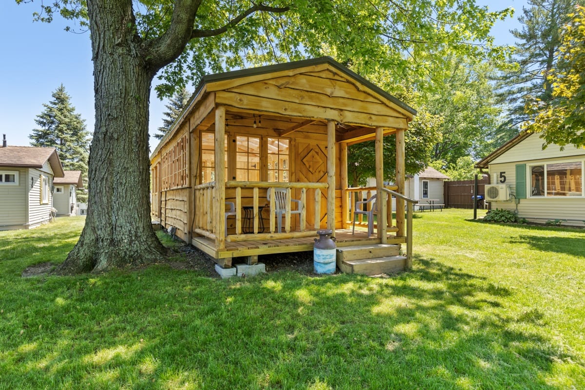 A charming wooden gazebo is nestled among lush green grass, offering a shaded seating area with ample natural light. Wooden railings frame the entrance, and large windows surround the structure, providing a view of the serene outdoor environment.