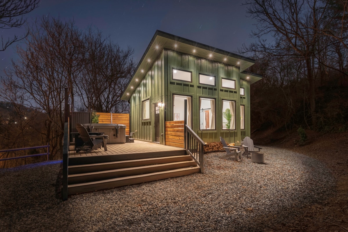 The cabin features a modern exterior with large windows and a green facade. A spacious deck with seating areas and a fire pit is highlighted, surrounded by gravel and trees, creating a serene setting. Soft lighting accentuates the structure against the night sky.