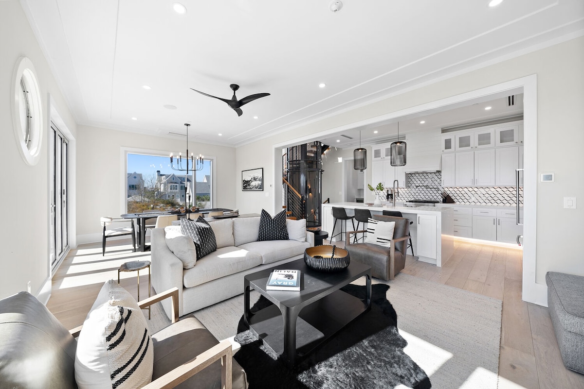 An open-concept living area features a spacious sofa and armchairs arranged around a dark coffee table. Natural light floods the space through large windows, illuminating the modern kitchen and dining area in the background. Contemporary decor and light-colored walls create a bright and airy ambiance.