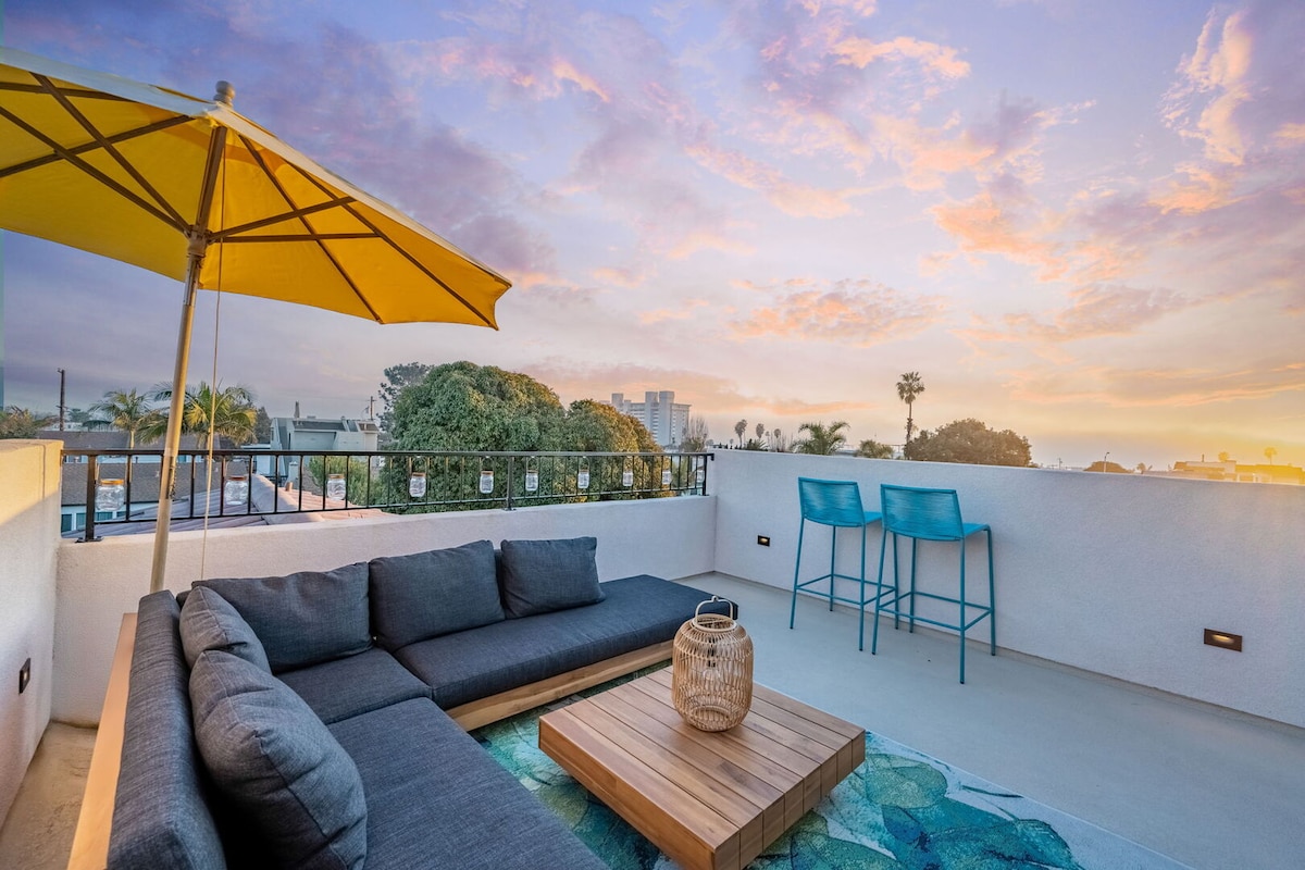 A spacious third-floor deck features a comfortable sectional sofa accompanied by a wooden coffee table. The area is shaded by a large yellow umbrella, with two blue bar stools positioned nearby. A colorful area rug adds a pop of color against the backdrop of a vibrant sunset sky.