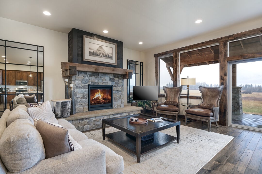 A living room showcases a stone fireplace framed by a wooden mantel, providing a warm focal point. A cozy sectional sofa and two leather chairs create a relaxed atmosphere, while a coffee table occupies the center of the room. Large windows allow natural light to enter.