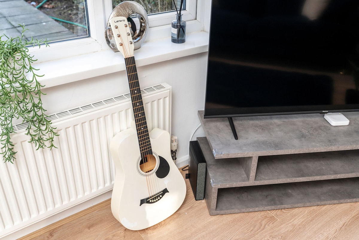 An acoustic guitar is positioned beside a radiator and a modern entertainment unit. The guitar, in a light color, leans against the wall, while the entertainment unit features a flat-screen TV and is made of gray tones, adding a touch of style to the room.
