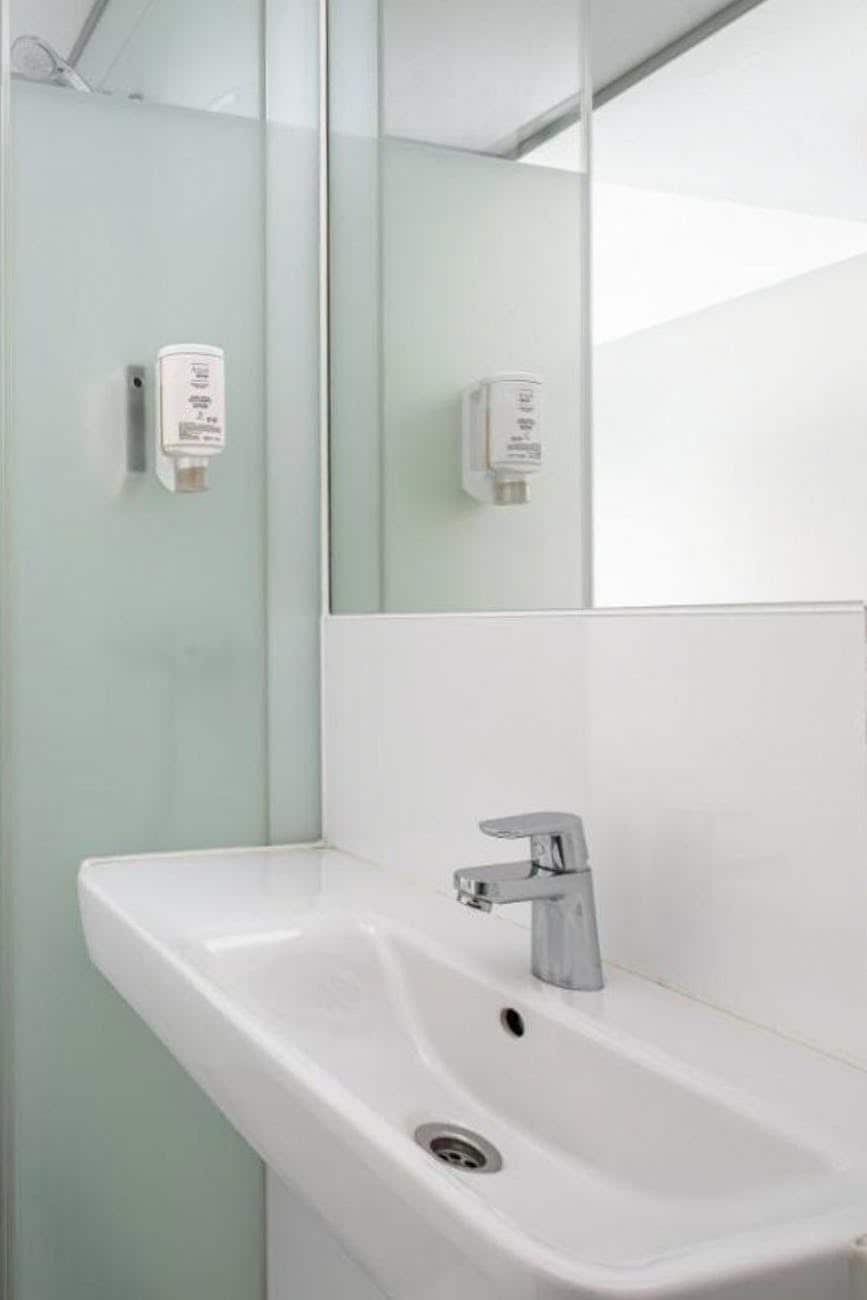A modern bathroom sink is shown with a sleek faucet, accompanied by wall-mounted dispensers for soap or lotion. A large mirror reflects the space, and frosted glass adds privacy while allowing light to filter in. The overall design emphasizes cleanliness and simplicity.