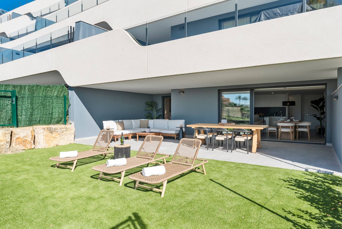 A private outdoor space features a lush green lawn accompanied by four sun loungers. A wooden dining table sits nearby, surrounded by chairs, while a cozy seating area is positioned against the wall. Large sliding glass doors lead into the interior living area.