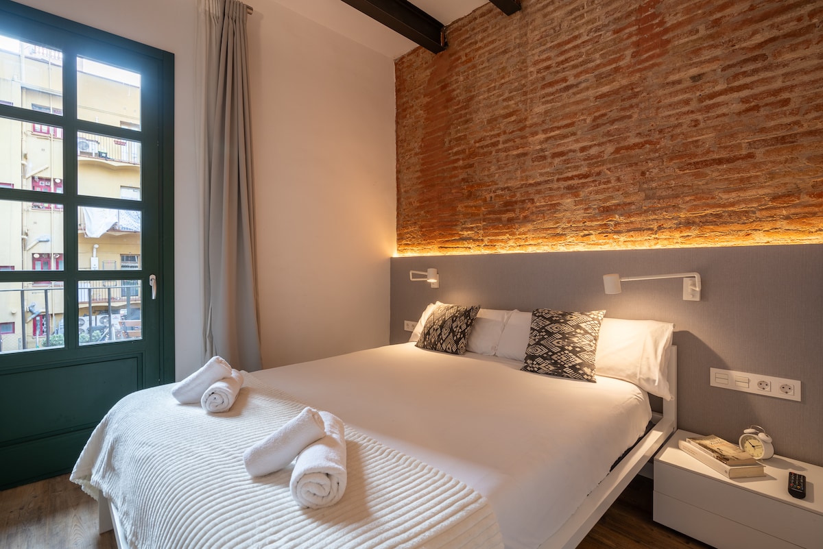 A bedroom features a spacious bed with neatly arranged white linens and towels. Exposed brick walls create a warm ambiance, complemented by soft lighting along the headboard. A green window allows natural light and offers a view of the outside area.