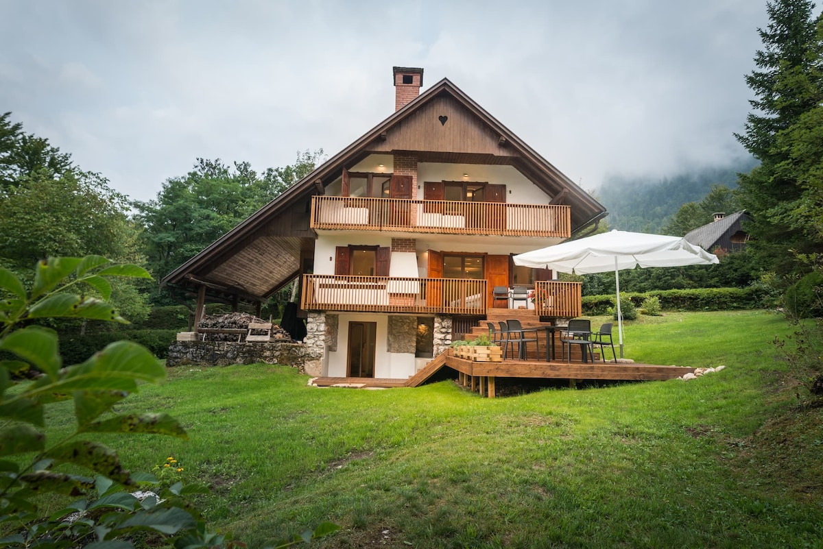 The two-story chalet is set in a green landscape, featuring a spacious deck with a table and chairs beneath a large umbrella. The wooden balcony showcases multiple levels, and the rustic design includes a stone foundation and grassy surroundings, with trees partially framing the view.