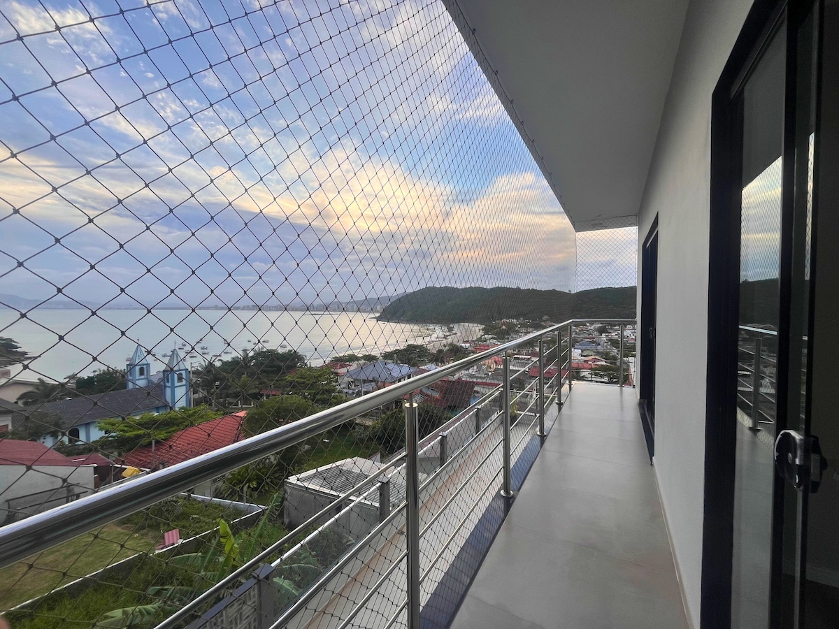 A balcony overlooks a coastal view, featuring a protective netting for safety. The landscape includes a glimpse of distant hills and a serene body of water. The flooring is smooth, and there are glass sliding doors leading inside.