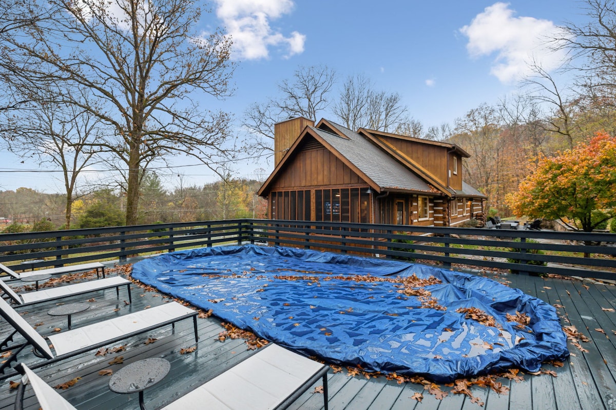 An expansive deck surrounds the pool area, featuring several white lounge chairs arranged neatly. The pool is covered with a blue tarp, showing a collection of fallen leaves. The cabin with wooden siding and surrounding trees completes the scenic outdoor view.