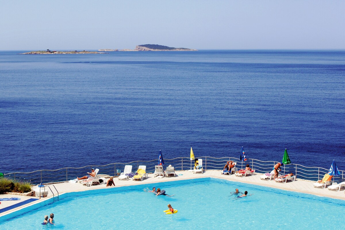 A vibrant swimming pool is framed by sun loungers in a variety of colors. The clear blue water reflects the expansive sea view, with an island visible in the distance. Relaxation spaces are situated around the pool area, inviting guests to enjoy the outdoor environment.